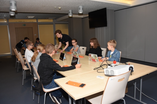 Zdjęcie - w pomieszczeniu, przy dużym podłużnym stole, siedzą dzieci biorące udział w zajęciach z programowania z użyciem systemu KANO. Przed nimi komputery z pomarańczowymi klawiaturami
